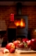 Red wine in bottle and glass with apple and strawberries on wooden table in front of roaring fire inside wood burning stove in brick fireplace