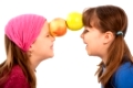 Laughing small girls facing each other, holding two apples together with forehead. 