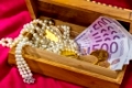 Gold in Münzen und Barren mit Schmuck auf rotem Samt. Symbolfoto für Reichtum, Luxus, Reichensteuer.