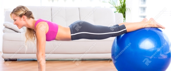 Slim blonde in plank position using exercise ball at home in the living room