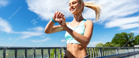 fitness, sport, people, technology and healthy lifestyle concept - smiling young woman with heart rate watch and earphones exercising outdoors