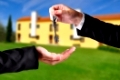 A hand giving a key to another hand. Both persons in suits. House in the background.