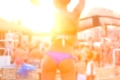 Sexy hot girl wearing brazilian bikini dancing on a beach party event in sunset. Crowd dancing and partying at poolside in background. Summer electronic music festival. Hot summer party vibe.