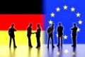 Modellfiguren, die Politiker symbolisieren, stehen vor den Flaggen von Deutschland und der EU. Zwei von Ihnen reichen sich die Hände.Digital Composite (DC)