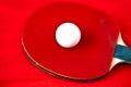 roter Tischtennisschlaeger mit Tischtennisball vor rotem Hintergrund