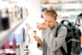 Blond young female traveler wearing coat and travel backpack choosing perfume in airport duty free store. Casual lady testing and buying cosmetics on the go in a beauty store.